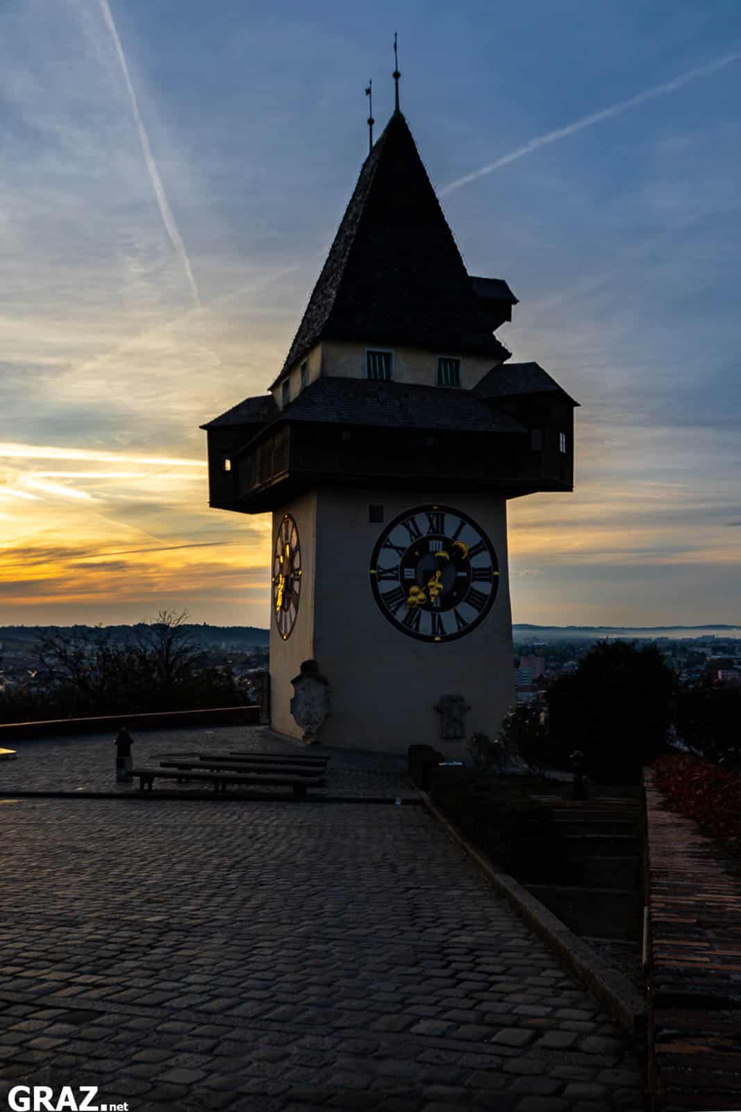 Grazer Uhrturm mit vorgelagertem Pflasterweg im Hochformat vor einem gelbblauen Morgenhimmel