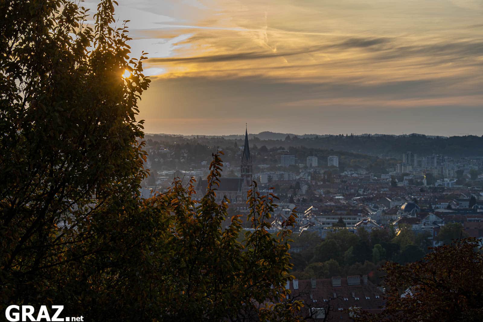 Blick vom Schloßberg auf Grazer Dächer und einen Kirchturm unter einem goldenen Morgenhimmel