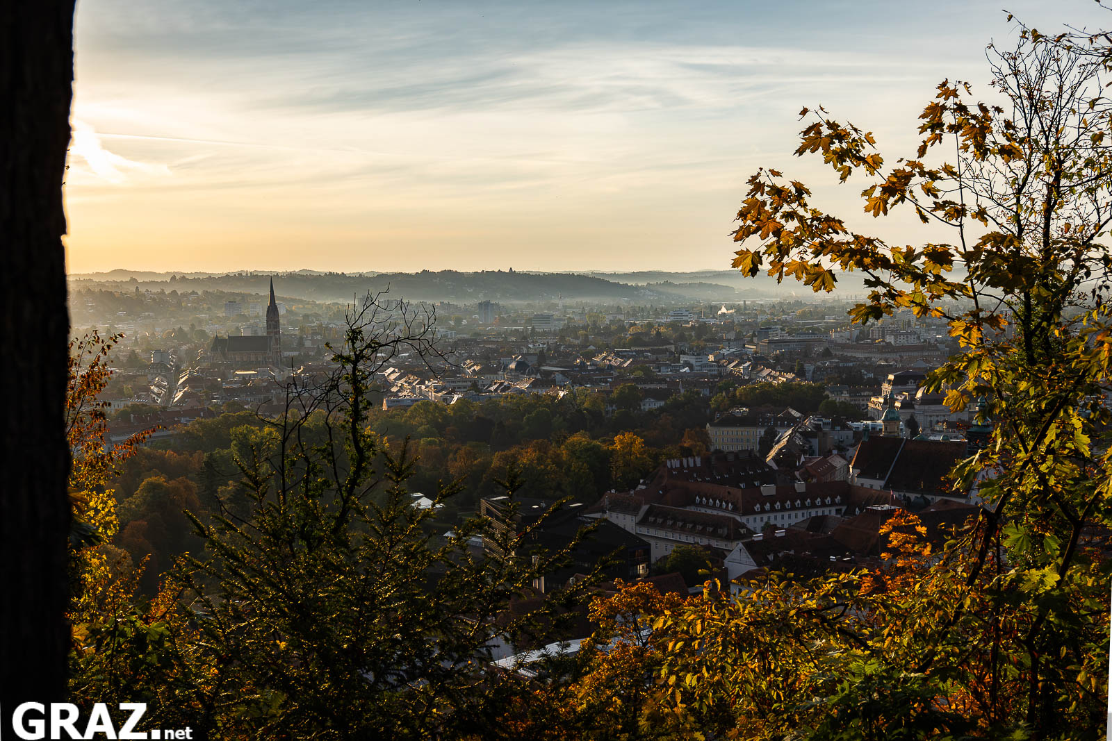 Panorama von Graz mit Kirchturm und dicht stehenden Häusern, eingerahmt von herbstlichen Bäumen