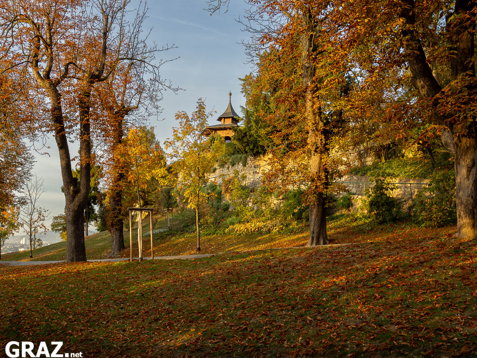 Orange und gelb gefärbte Bäume am Grazer Schloßberg mit einem Pavillon oberhalb des Hangs