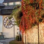Grazer Uhrturm mit goldenem Uhrzeiger neben einer Mauer mit roten und orangefarbenen Herbstblättern