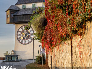 Grazer Uhrturm mit goldenem Uhrzeiger neben einer Mauer mit roten und orangefarbenen Herbstblättern
