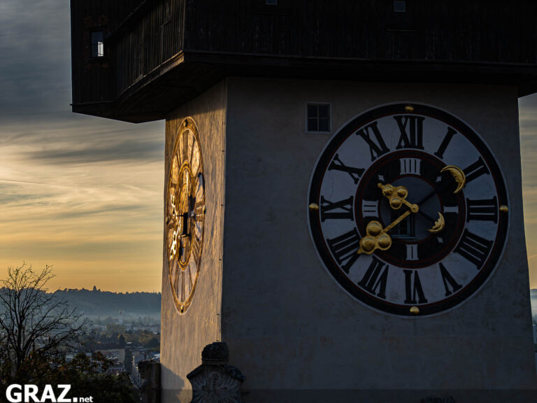 Nahansicht des Zifferblatts und des hölzernen Umgangs am Grazer Uhrturm vor einem herbstlichen Morgenhimmel
