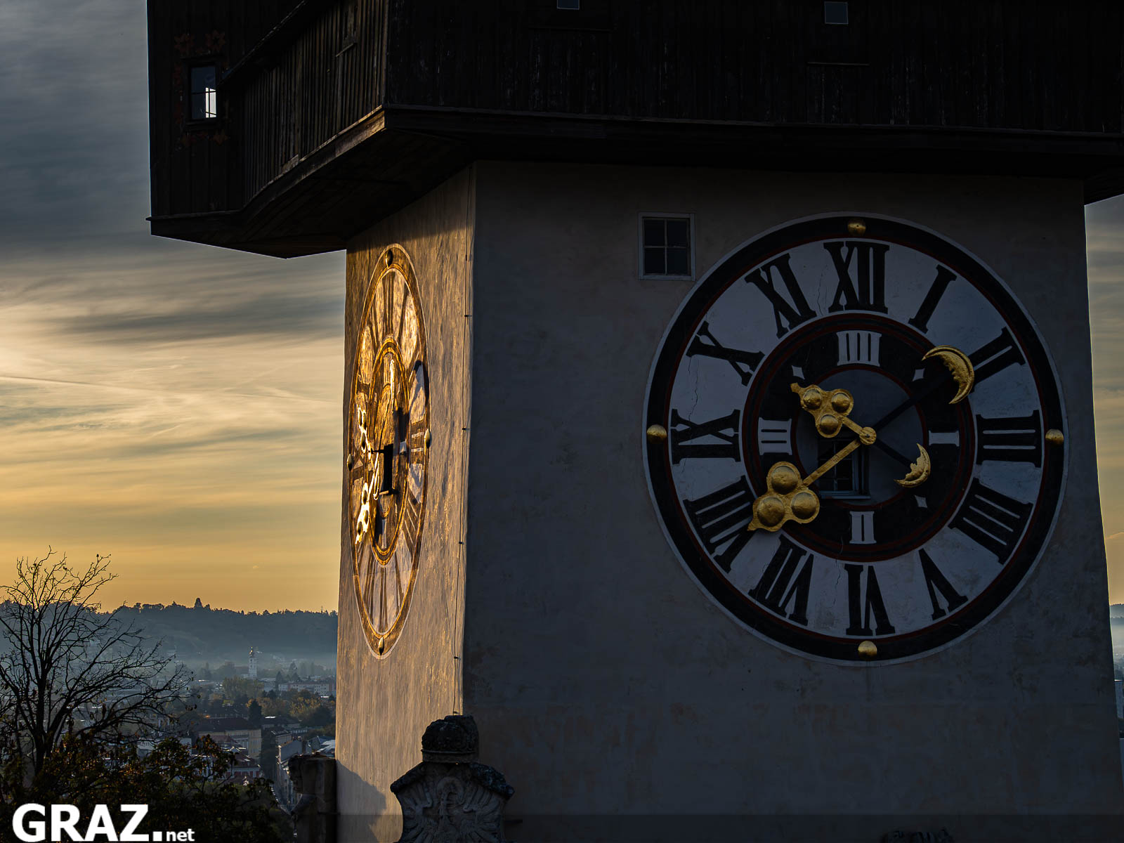 Nahansicht des Zifferblatts und des hölzernen Umgangs am Grazer Uhrturm vor einem herbstlichen Morgenhimmel