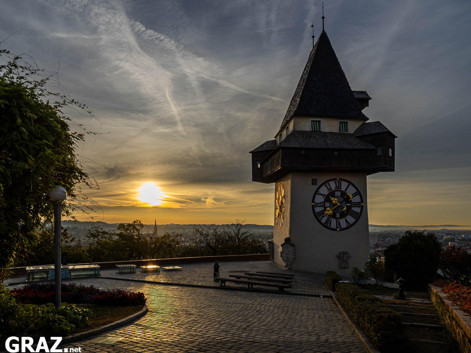 Aufgehende Sonne links neben dem Grazer Uhrturm mit goldenem Licht auf dem gepflasterten Platz