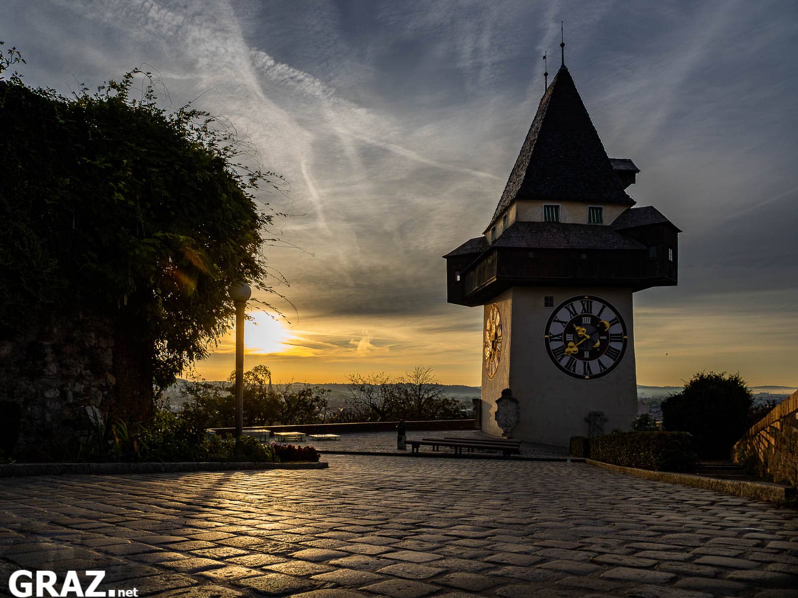 Grazer Uhrturm über einem im Morgenlicht glänzenden Pflasterplatz unter einem Himmel mit feinen Wolken