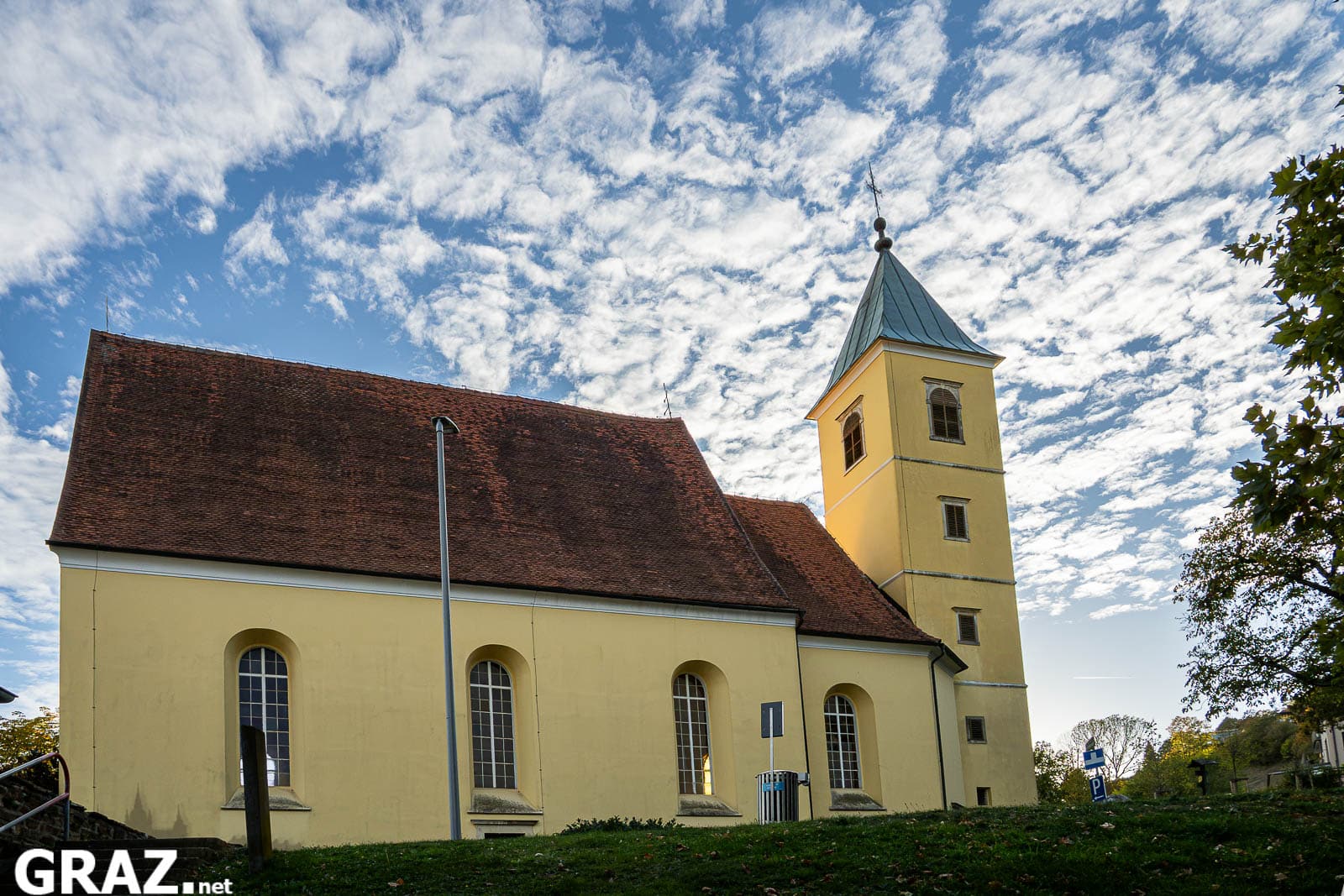 Gelbe Schlosskirche St. Martin in Graz mit rotem Ziegeldach unter einem Himmel mit weißen Wolken