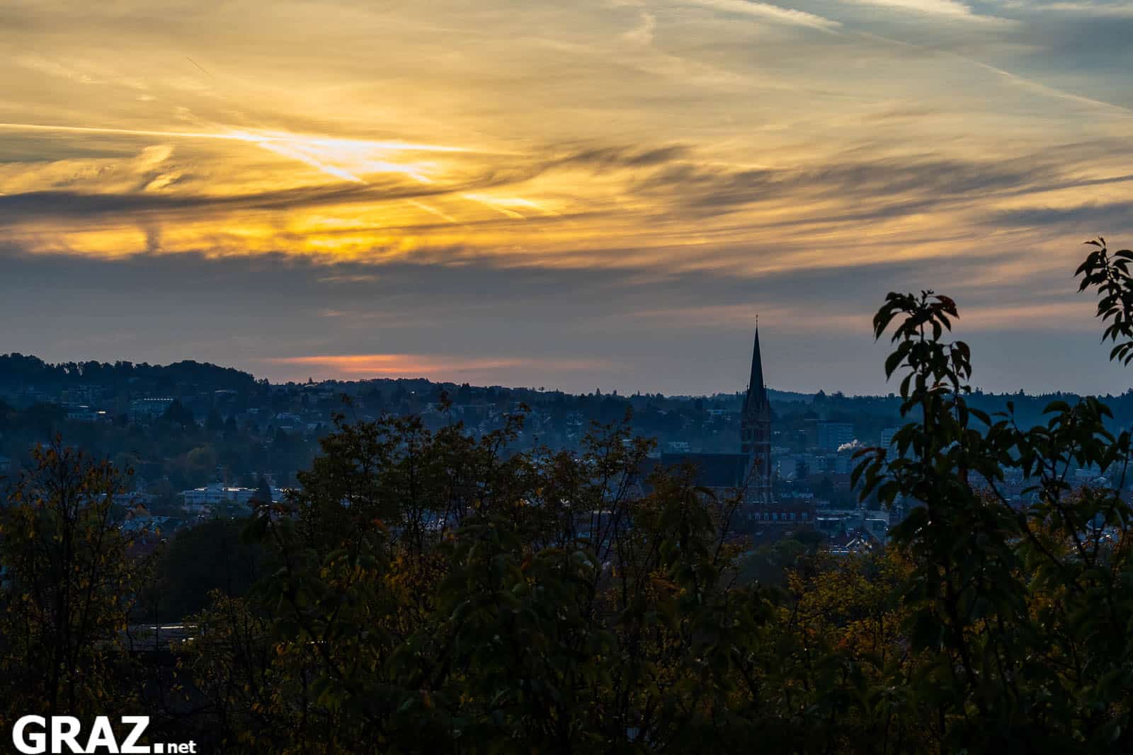 Grazer Dächer und ein schlanker Kirchturm unter einem orangegelben Morgenhimmel, davor dunkle Baumkronen