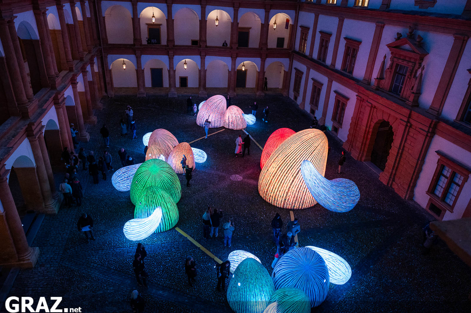 Blick aus den Arkaden auf die bunt leuchtenden „Whispering Mountains“ von ENESS und die Gäste im Schlosshof.