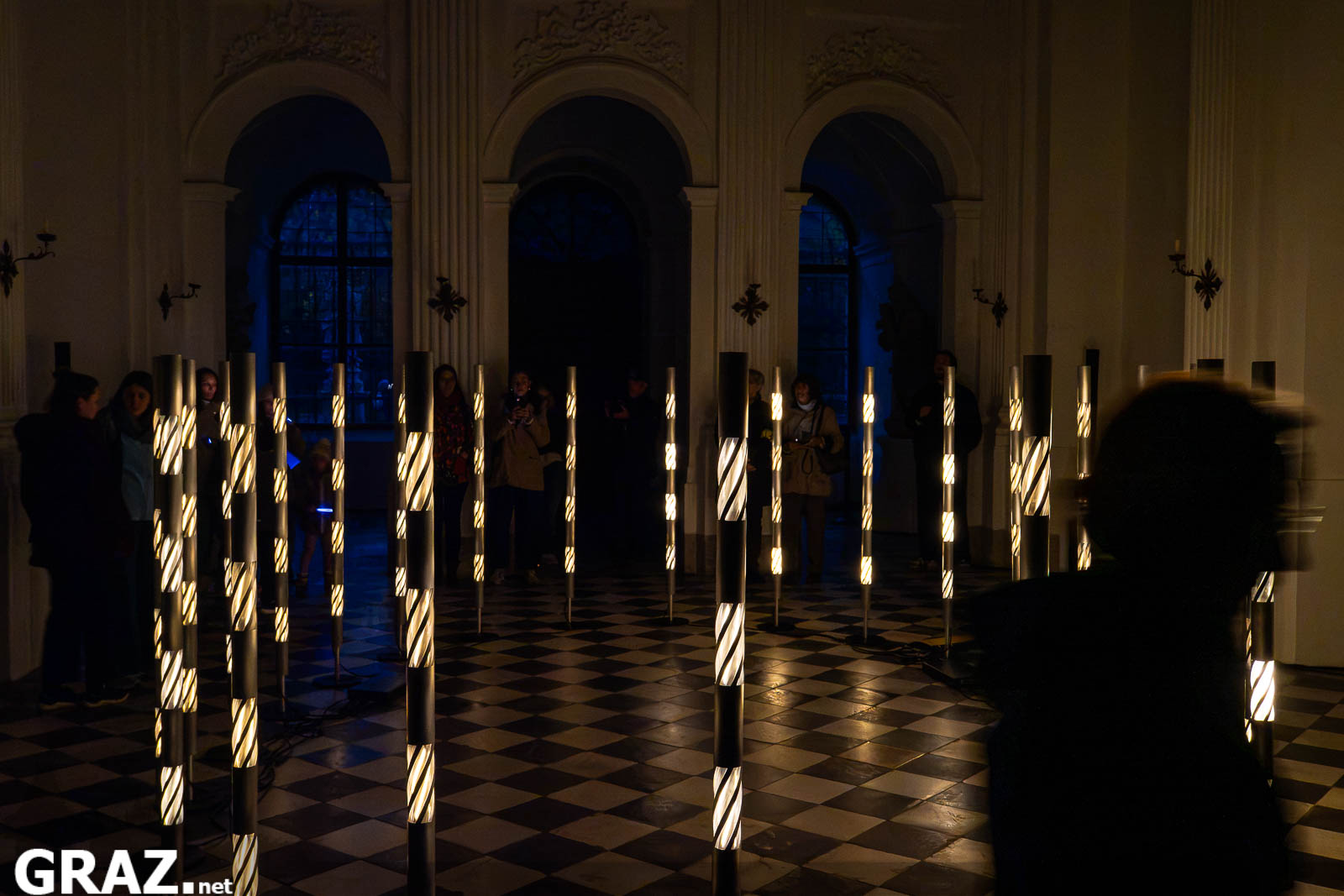 Leuchtstäbe von Robin Beekmans „Continuum“ in der Schlosskapelle Eggenberg, rechts eine Person in Bewegung.