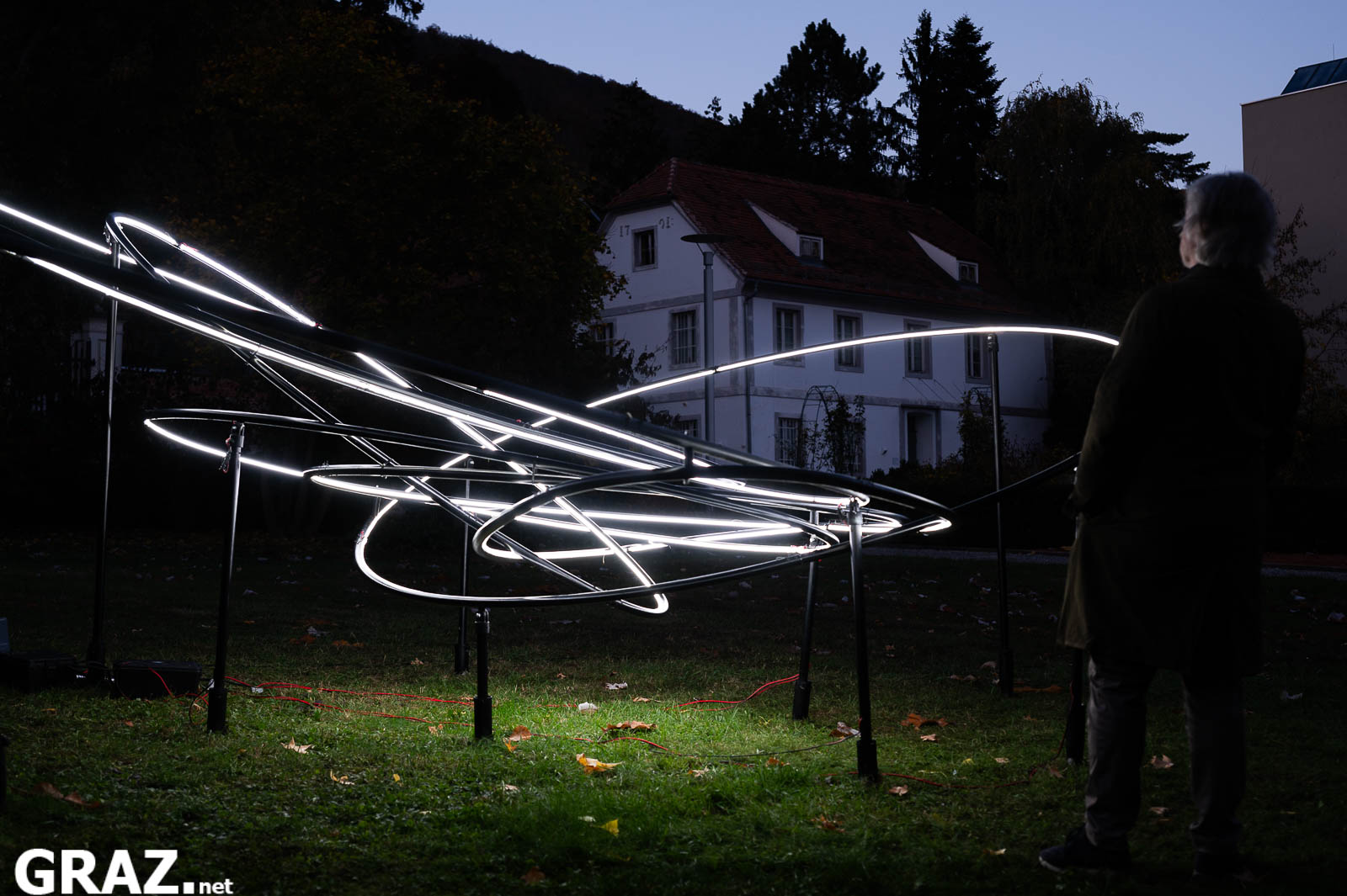 Eine Person betrachtet Miriam Hamanns „Planet 9“ mit verschlungenen weißen Lichtbahnen im Schlosspark Eggenberg.
