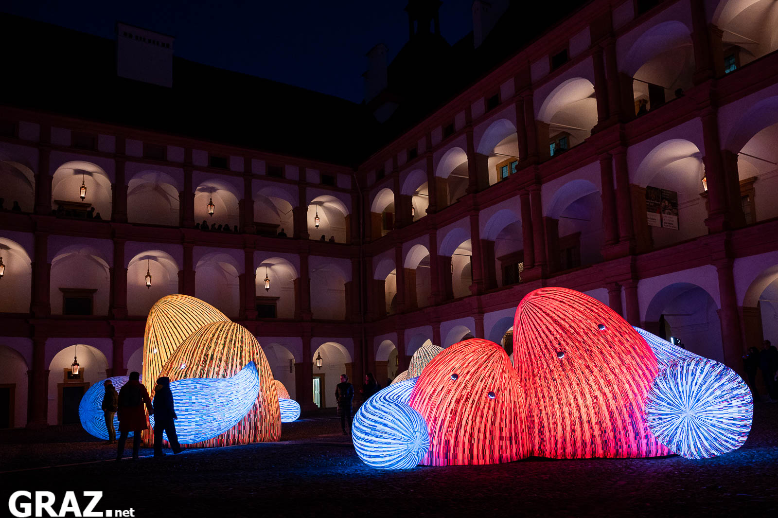 Gäste zwischen rot und gelb leuchtenden „Whispering Mountains“ von ENESS vor den Arkaden von Schloss Eggenberg.