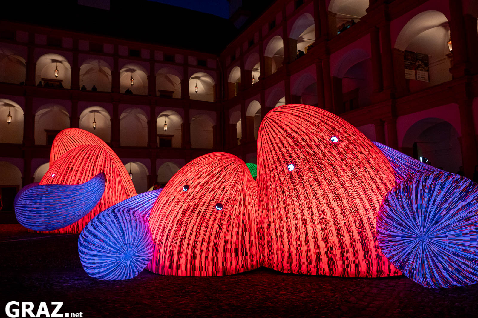 Rot leuchtende Bergfiguren mit blau gestreiften Armen aus ENESS’ „Whispering Mountains“ im Innenhof von Schloss Eggenberg.