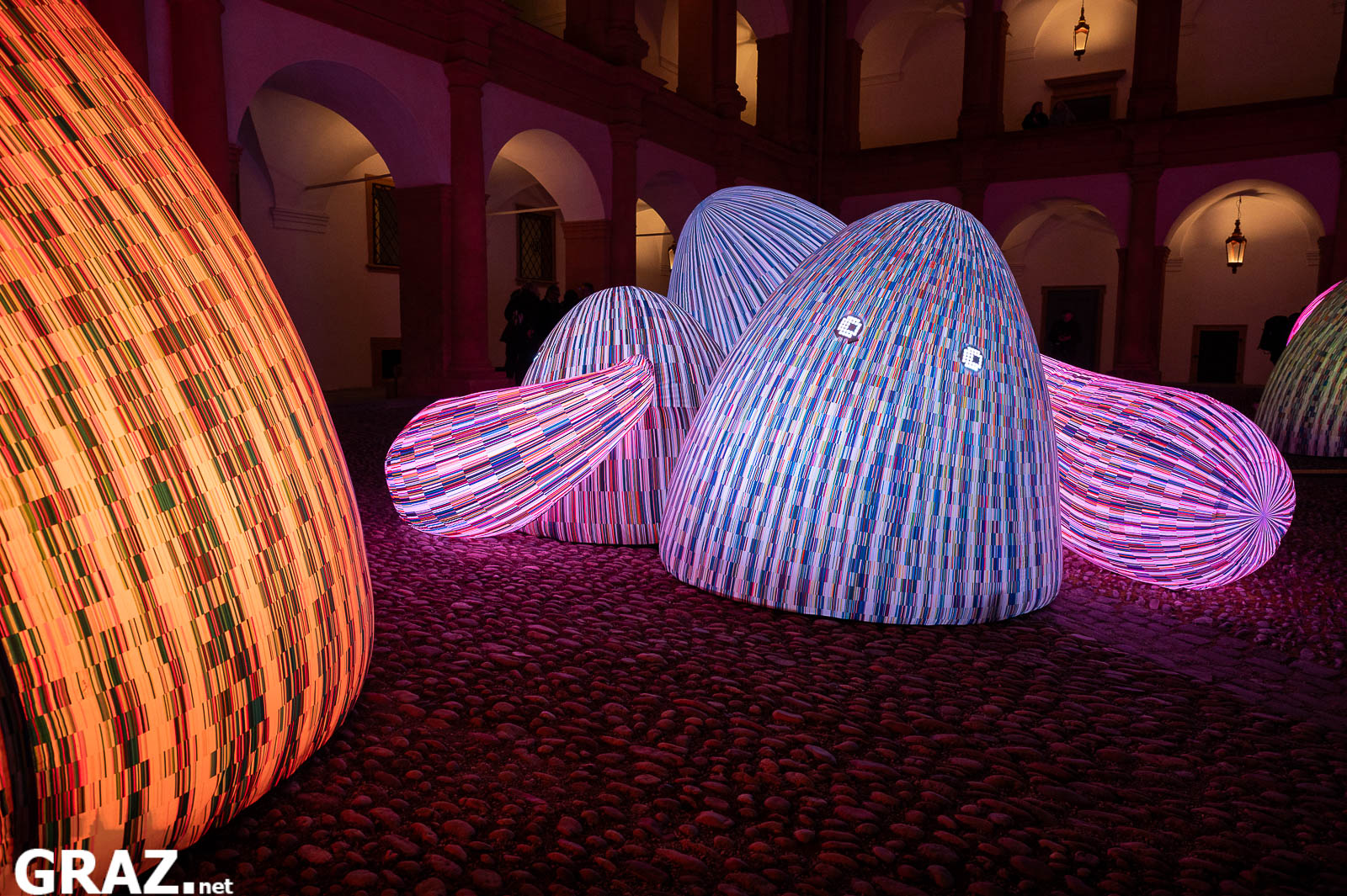 Eine blau und rosa gestreifte Bergfigur aus „Whispering Mountains“ von ENESS mit kleinen leuchtenden Augen im Schlosshof.