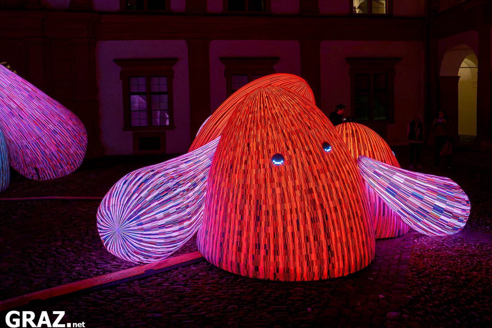 Eine rot leuchtende Bergfigur mit ausgebreiteten gestreiften Armen aus ENESS’ „Whispering Mountains“ im Schlosshof.