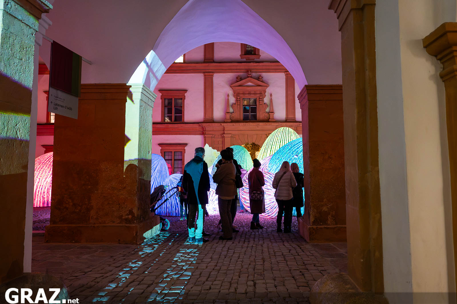 Blick durch einen Torbogen auf Gäste und die bunt leuchtenden „Whispering Mountains“ von ENESS in Schloss Eggenberg.
