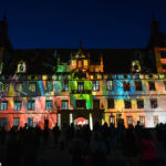 Die gesamte Schlossfassade erstrahlt in bunten Projektionen von „Kadenzen“ von Daniel Rossa, mit Gästen im Vordergrund.