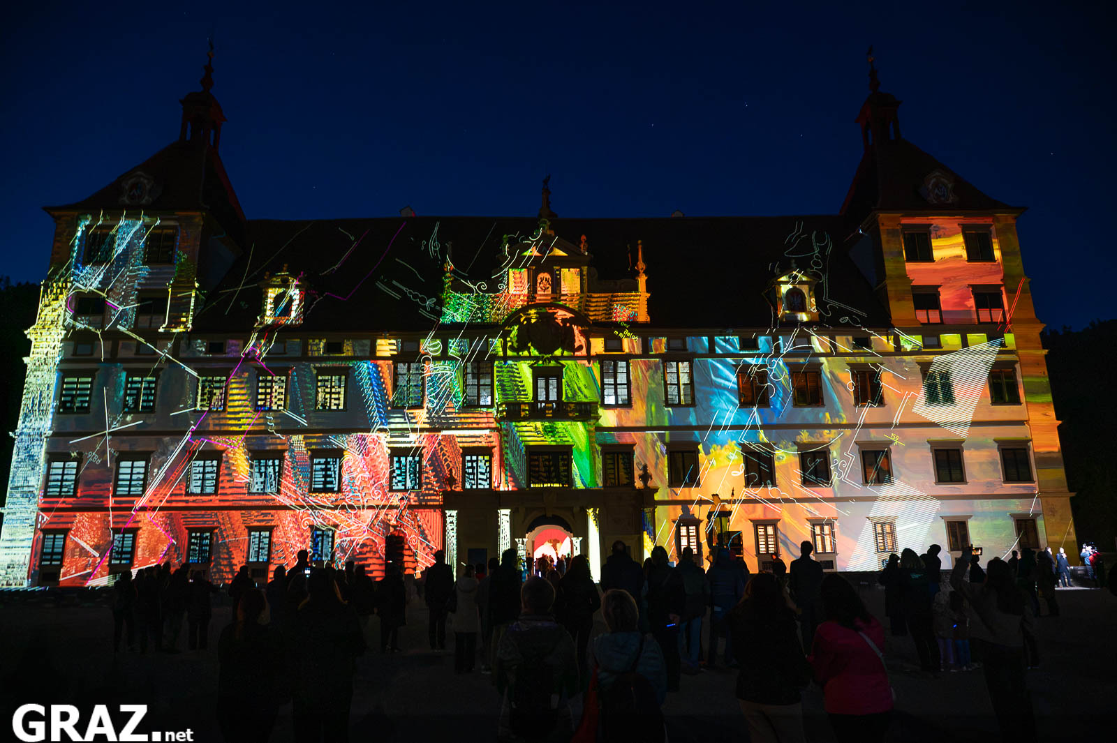 Die gesamte Schlossfassade erstrahlt in bunten Projektionen von „Kadenzen“ von Daniel Rossa, mit Gästen im Vordergrund.