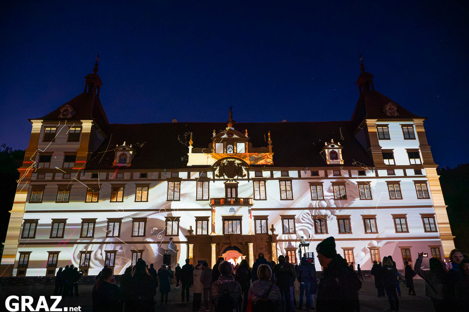 Weiße Projektionsflächen aus „Kadenzen“ von Daniel Rossa auf der Fassade von Schloss Eggenberg vor dunklem Abendhimmel.