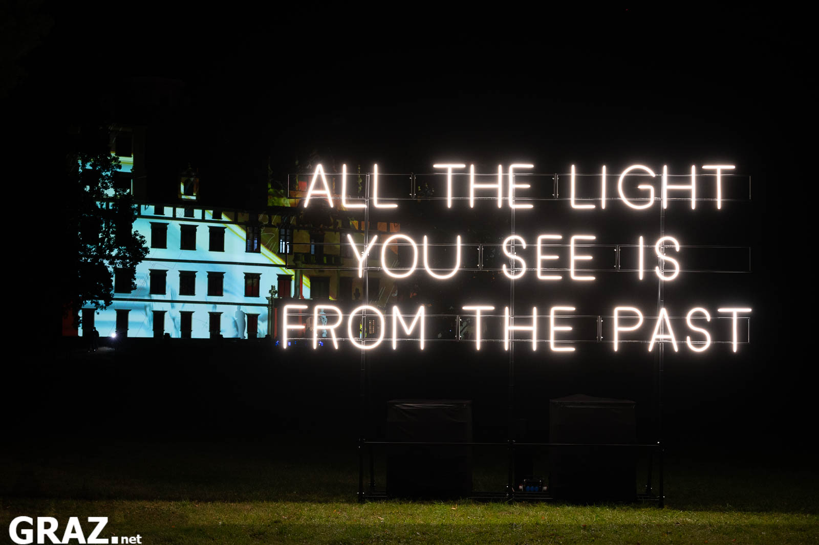 Die Leuchtschrift „All the light you see“ von Alicia Eggert im Schlosspark mit einem beleuchteten Gebäude im Hintergrund.