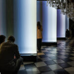 Zwei Gäste sitzen vor den farbigen Lichttafeln von Inge Dicks „jahres licht weiss“ im Planetensaal von Schloss Eggenberg.