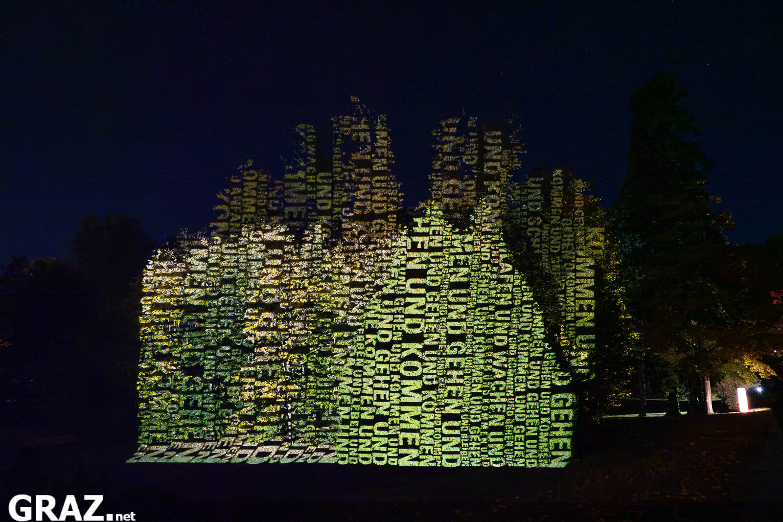 Gelbe und grüne Wörter aus „Räume der Zeit“ von Hartung & Trenz werden auf Baumkronen im Schlosspark Eggenberg projiziert.